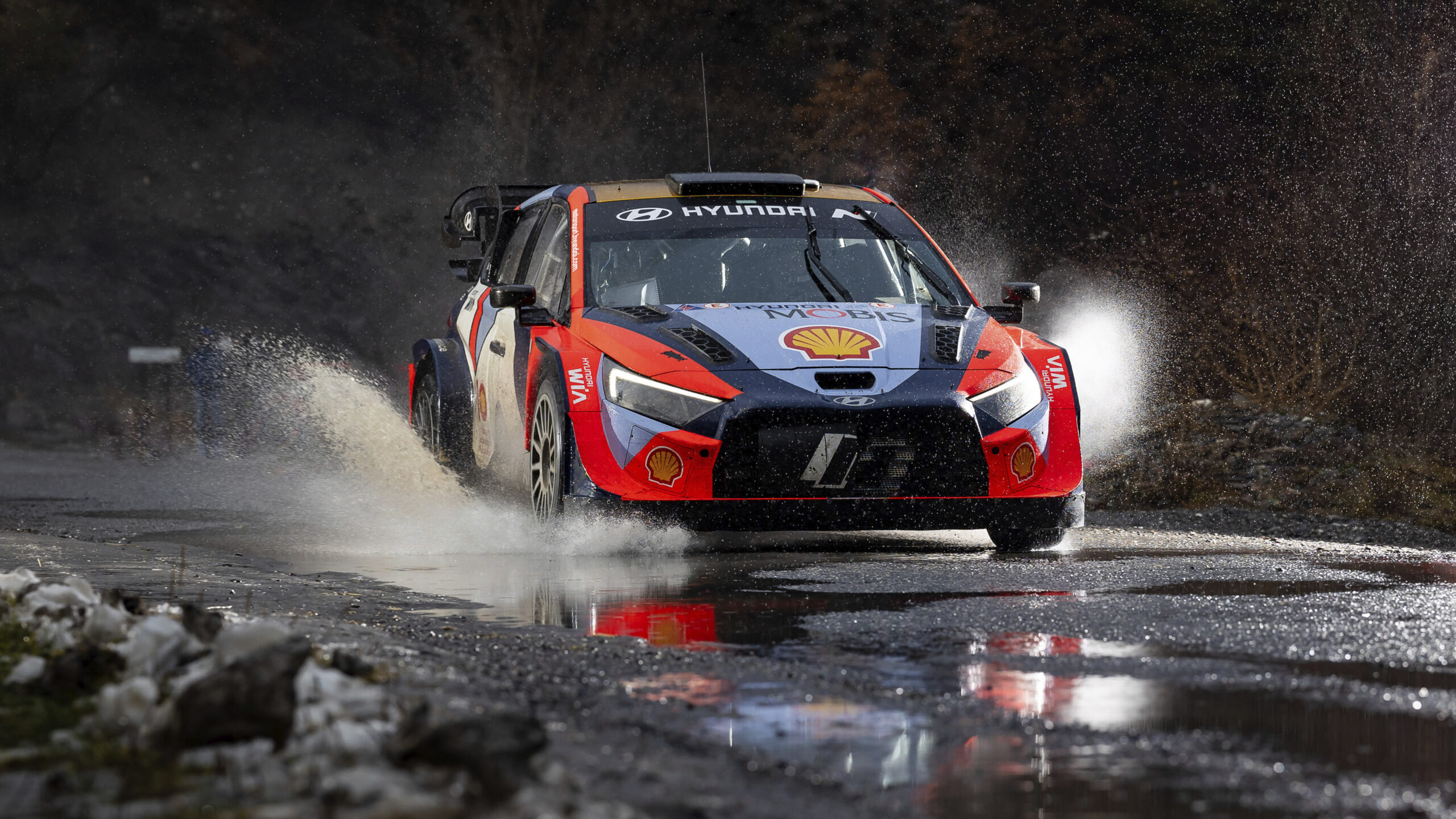 A rally car at speed through standing water on a night stage